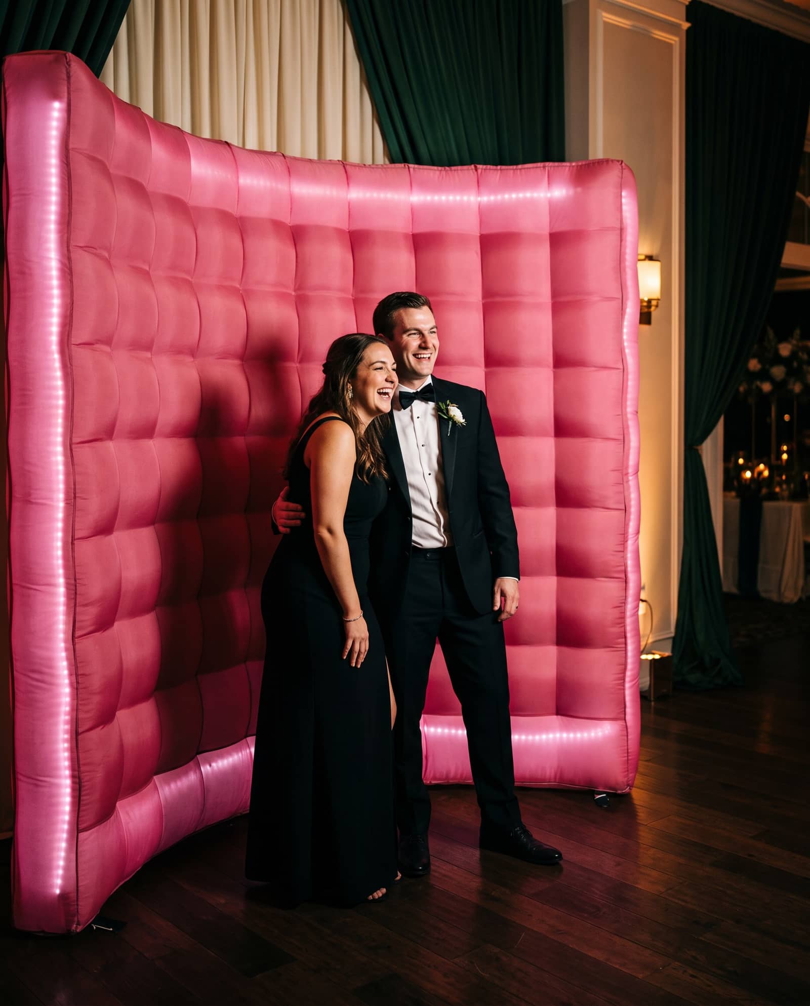 A couple laughing in front of a ten foot LED inflatable photo booth wall lit deep rose pink at an evening wedding reception